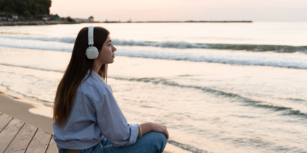 Young woman wearing white headphones sitting on a wooden boardwalk, looking out at the calm ocean waves at sunset.
